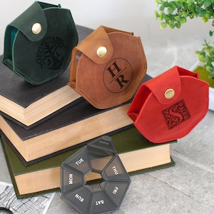 Può includere: Tre astucci per pillole in pelle verde, marrone e rossa, ciascuno con un design in rilievo unico e una chiusura a scatto dorata. Un organizer settimanale nero per pillole è in primo piano, con una pila di libri sullo sfondo.