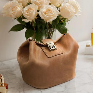 Pode incluir: Uma bolsa de mão de couro marrom claro com um fecho dourado e as iniciais "VS" gravadas na frente.