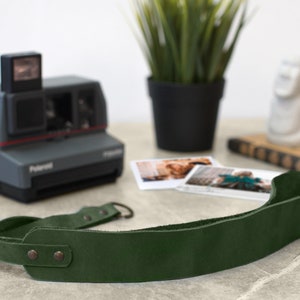 May include: A dark green leather camera strap with two metal snaps and a metal loop. The strap is lying on a light gray surface.
