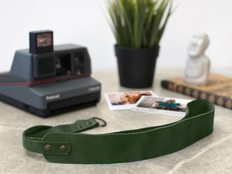 May include: A dark green leather camera strap with two metal snaps and a metal loop. The strap is lying on a light gray surface.