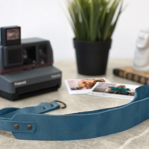 May include: A blue leather camera strap with two metal snaps and a metal loop. The strap is lying on a light gray surface.