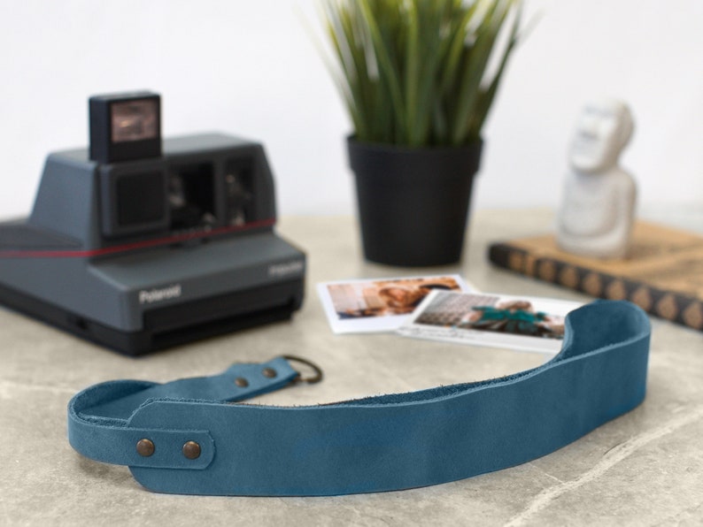 May include: A blue leather camera strap with two metal snaps and a metal loop. The strap is lying on a light gray surface.