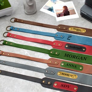 May include: Eight personalized leather camera straps with metal rings and gold-toned accents. The straps are in various colors, including brown, blue, red, green, gray, and black. Each strap has a personalized nameplate with a different name.