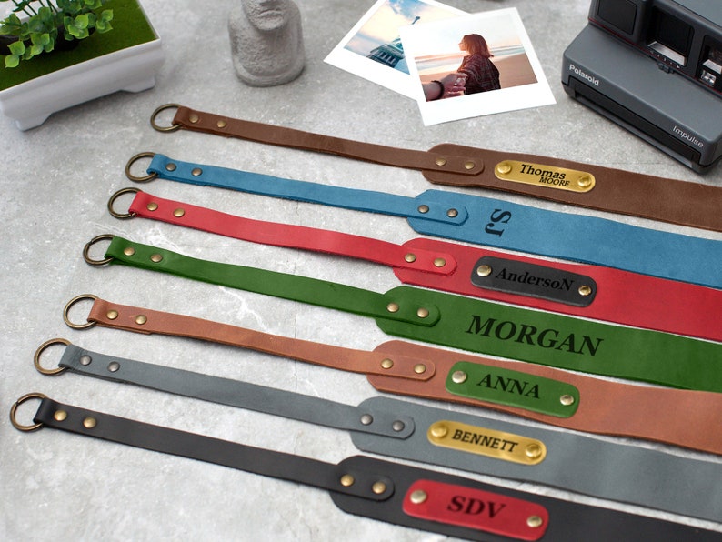May include: Eight personalized leather camera straps with metal rings and gold-toned accents. The straps are in various colors, including brown, blue, red, green, gray, and black. Each strap has a personalized nameplate with a different name.