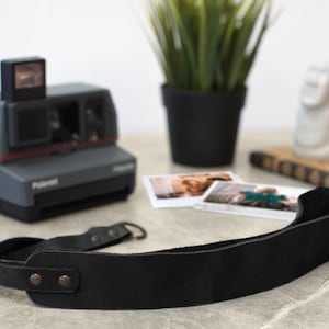 May include: A black leather camera strap with two metal rivets. The strap is lying on a table next to a vintage Polaroid camera.