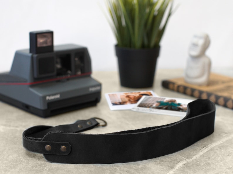 May include: A black leather camera strap with two metal rivets. The strap is lying on a table next to a vintage Polaroid camera.