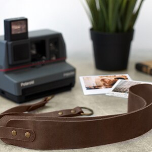 May include: A brown leather camera strap with metal hardware and two brass rivets. The strap is lying on a light gray surface.