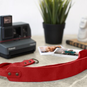 May include: Red leather camera strap with metal hardware. The strap is lying on a table with a vintage Polaroid camera and some photos.