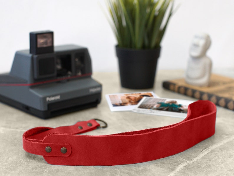 May include: Red leather camera strap with metal hardware. The strap is lying on a table with a vintage Polaroid camera and some photos.