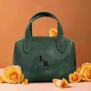 May include: A green leather handbag with two handles, featuring the initials "L R" in black. The bag is surrounded by yellow roses. The background is a warm orange colour.