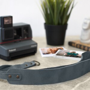 May include: A gray leather camera strap with two metal snaps and a loop on one end. The strap is lying on a gray surface.