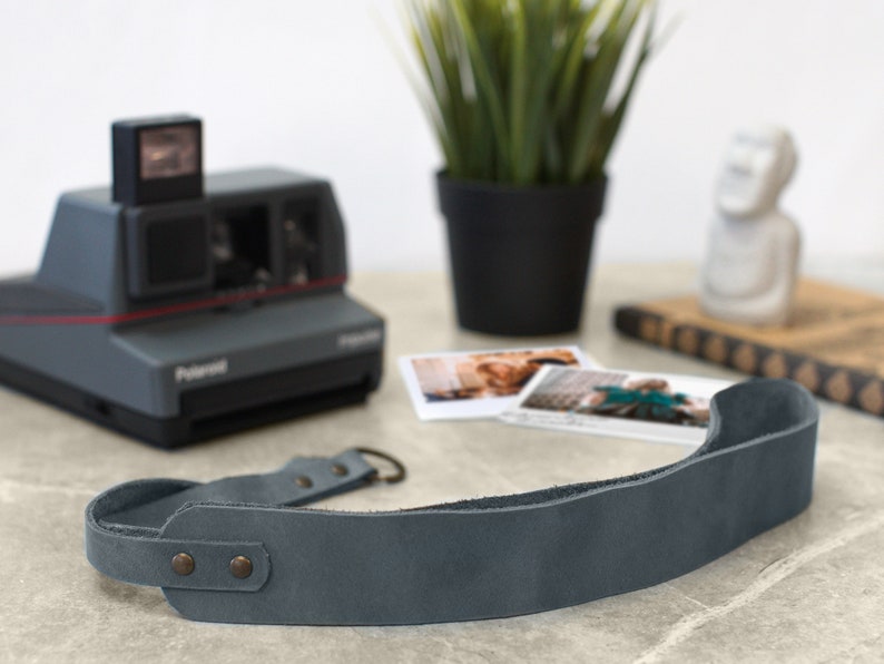 May include: A gray leather camera strap with two metal snaps and a loop on one end. The strap is lying on a gray surface.
