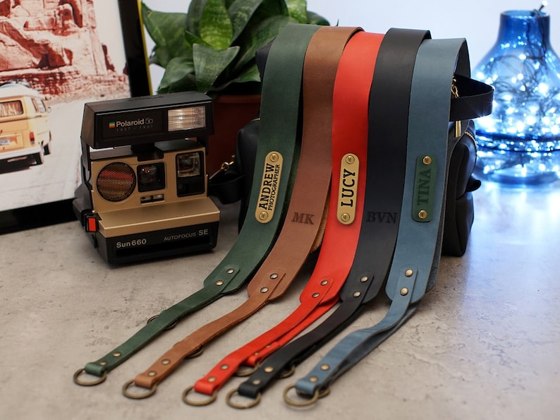 May include: Five different colored leather camera straps with personalized brass name tags. The straps are green, brown, red, black, and blue. The name tags read "Andrew Photographer", "MK", "Lucy", "BVM", and "Tina".