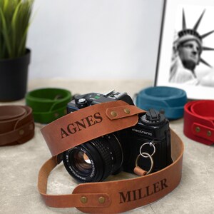 May include: A brown leather camera strap with the names "AGNES MILLER" engraved on it. The strap is attached to a black camera with a silver lens.