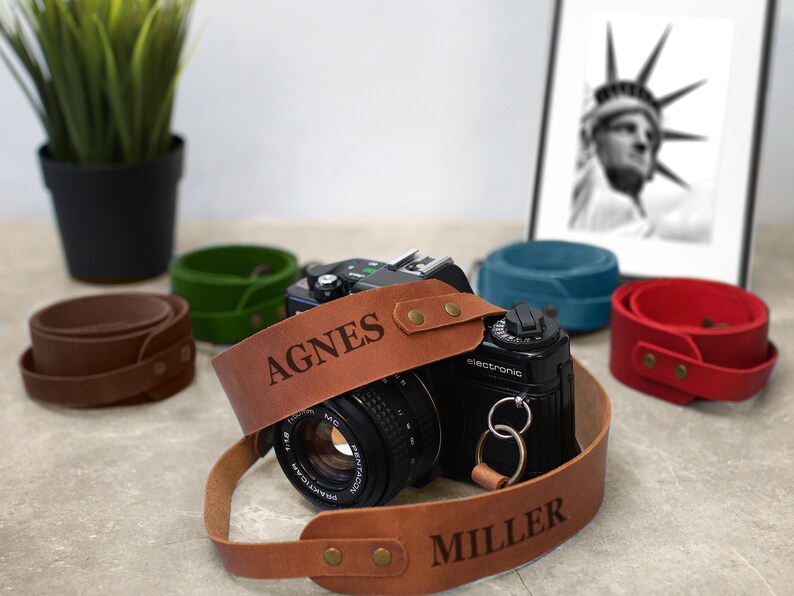 May include: A brown leather camera strap with the names "AGNES MILLER" engraved on it. The strap is attached to a black camera with a silver lens.