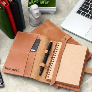 May include: A brown leather notebook cover with a pen holder, a card slot, and the name "Bennett" embossed on the front. The cover is open to reveal a spiral bound notebook with blank pages.