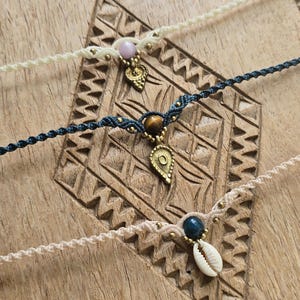 Peut inclure: Gros plan sur quatre bracelets macramé faits à la main avec des perles et des breloques décoratives. Les bracelets sont de couleur crème, marine et beige, chacun avec un charme unique, dont un coquillage, une pierre précieuse et un pendentif en forme de feuille.
