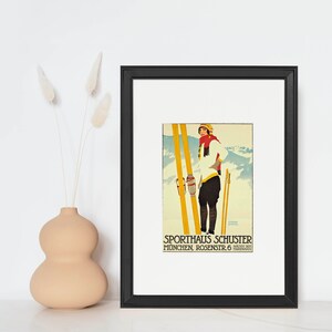 Op de afbeelding: Een vintage skiposter met een vrouw in een wit skipak en rode sjaal die op ski's staat tegen een besneeuwde bergachtergrond. De poster is zwart ingelijst en heeft de tekst "SPORTHAUS SCHUSTER MUNCHEN ROSENSTR. 6 NACHST DER HARDENBR&Uuml;CKE".
