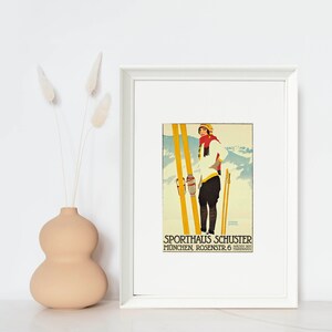 Op de afbeelding: Een vintage poster die reclame maakt voor Sporthaus Schuster in M&uuml;nchen, Duitsland. De poster toont een ski&euml;r in een witte trui, rode sjaal en zwarte broek die op gele ski's staat tegen een besneeuwde bergachtergrond. De tekst luidt "Sporthaus Schuster Munchen Rosenstr. 6 Nachst dem Hauptbahnhof".