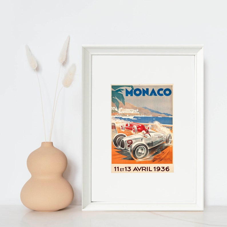 May include: Vintage poster advertising the Monaco Grand Prix, featuring a silver race car with a red car in the background. The poster is framed in white and has the text "MONACO" and "11 et 13 AVRIL 1936".