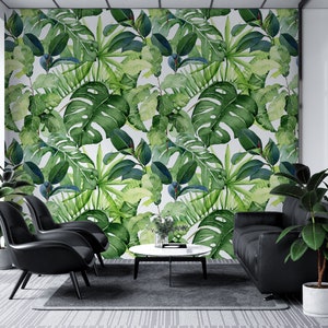 Op de afbeelding: Een woonkamer interieur met een witte muur bedekt met een tropische bladbehang. De kamer heeft twee zwarte stoelen, een zwarte bank en een ronde salontafel.