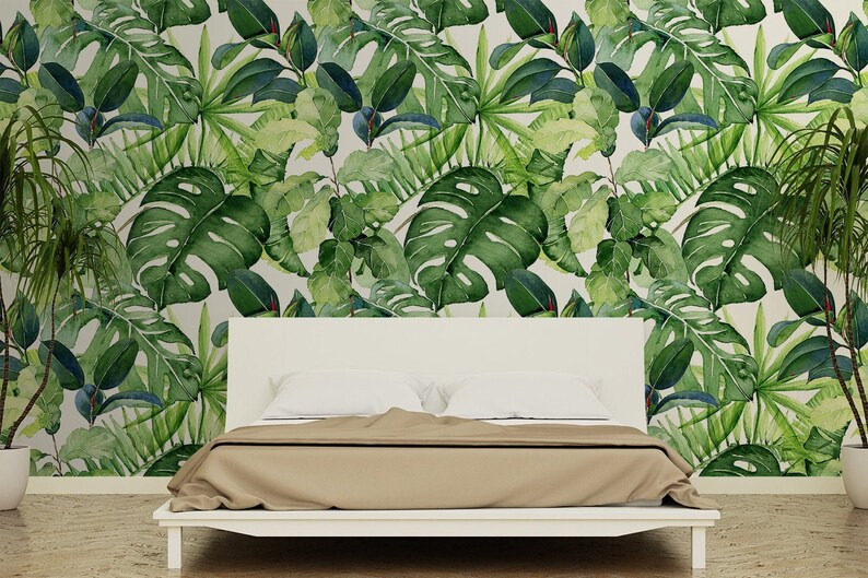 Op de afbeelding: Een wit bedframe met een beige hoofdeinde en een tan dekbedovertrek. Het bed staat voor een muur met een tropisch bladmotief behang.