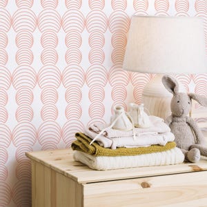 May include: A white dresser with a pink and white geometric patterned wallpaper. On top of the dresser are a lamp, a stack of folded clothes, and a stuffed bunny.