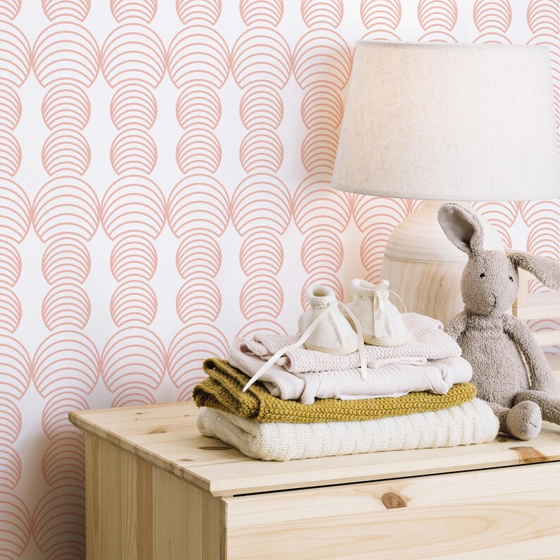 May include: A white dresser with a pink and white geometric patterned wallpaper. On top of the dresser are a lamp, a stack of folded clothes, and a stuffed bunny.