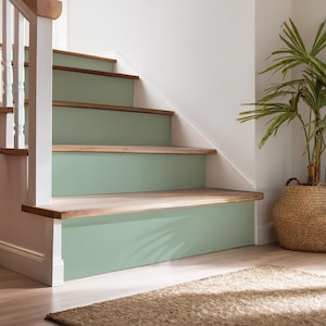 Op de afbeelding: Een set houten trappen geverfd in een lichtgroene kleur. De trappen bevinden zich in een huis met witte muren en een houten vloer. Een geweven mand met een groene plant staat in de hoek van de kamer.