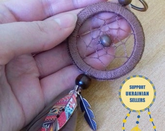 May include: Brown dream catcher keychain with a beaded tassel and a colorful feather. The keychain is held in a hand. The text "Support Ukrainian Sellers" is visible in the background.