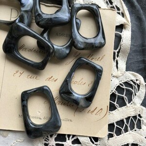 May include: A collection of dark gray and white marbled resin rings with rectangular openings. The rings are scattered on a vintage paper with handwritten text and a lace trim.