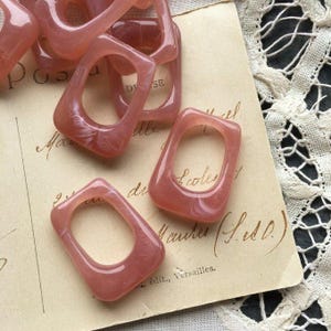 May include: Several translucent, rectangular pink resin rings with rounded corners are scattered on a vintage paper background. The rings have a hollow center. The paper has handwritten script and is next to a white lace doily.