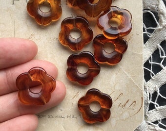 May include: A collection of eight brown plastic flower-shaped beads with a hole in the center. The beads are arranged on a white surface with a lace overlay.