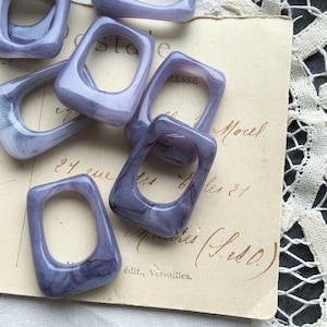 May include: Several translucent, rectangular lavender-colored beads with rounded corners are scattered across a vintage paper. The beads have a marbled effect, with darker purple swirls within the lighter lavender. The paper has handwritten text and a lace border.