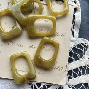 May include: A collection of olive-green, rectangular resin rings with rounded corners and a central opening. The rings have a marbled appearance and are arranged on a vintage paper with handwritten text. The rings are likely for jewelry making or crafting.