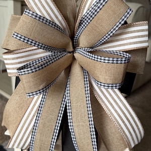 May include: A large decorative bow featuring burlap, black and white gingham, and striped ribbons. The bow is a mix of textures and patterns, perfect for adding a rustic touch to home decor or gift wrapping.
