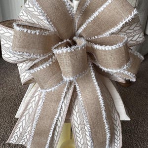May include: A large, brown burlap bow with white frayed edges and a white braided pattern. The bow is tied around a white lantern.