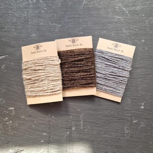 Op de afbeelding: Drie spoelen garen in verschillende kleuren: beige, bruin en grijs. Elke spoel is gelabeld met "Gosh Darn It" en heeft een kleine illustratie van een naald en draad.