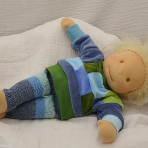 Könnte beinhalten: Eine handgemachte Puppe mit blonden Haaren, die ein blau-grünes gestreiftes Hemd und eine blau-graue gestreifte Hose trägt, liegt auf einer weißen Decke.