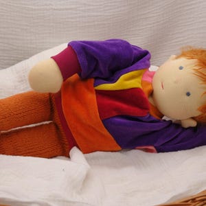 Könnte beinhalten: Eine handgefertigte Puppe mit orangefarbener und lilafarbener Kleidung liegt in einem Weidenkorb. Die Puppe hat einen hellen Teint und rote Haare.