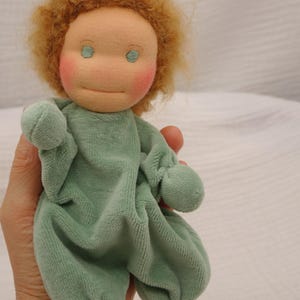 Könnte beinhalten: Eine handgefertigte Puppe mit einem weichen, plüschigen Körper in Hellgrün. Die Puppe hat ein pfirsichfarbenes Gesicht mit rosigen Wangen, gestickten Augen und hellbraunem, lockigem Haar. Die Arme und Beine der Puppe sind abgerundet.