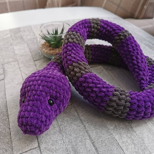 May include: A purple and grey crocheted snake toy. The snake is curled up in a circle with its head facing the camera. The snake has black eyes and a textured surface.