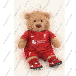May include: A plush teddy bear wearing a red soccer uniform with the Liverpool Football Club logo and the Standard Chartered sponsor. The bear is wearing red shorts, shoes, and a jersey. The background has watercolor paint splatters.