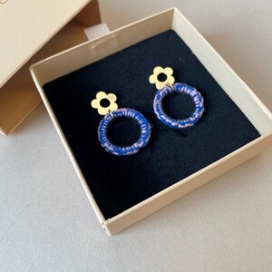 May include: A pair of blue and pink woven hoop earrings with gold flower charms. The earrings are in a small, beige box.