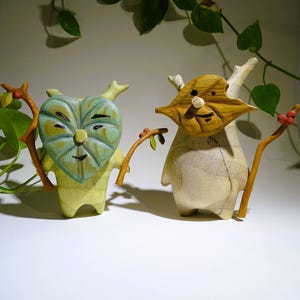 Könnte beinhalten: Zwei Holzfiguren mit einzigartigen Designs. Eine hat ein herzförmiges Gesicht mit blau-grünen Akzenten, die andere ein maskenartiges Gesicht. Beide halten Zweige mit Beeren und Blättern. Sie wirken handgefertigt und haben einen verspielten Charakter.