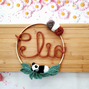 Peut inclure: Un cercle en bois avec le nom "Elia" écrit en fil brun. Un panda en crochet est couché sur des feuilles vertes sous le nom. Un pompon brun et blanc est attaché au cercle.