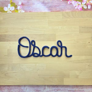 May include: A dark blue knitted cord shaped into the cursive name Oscar. The letters are formed from a single continuous piece of yarn with a textured, woven appearance, creating a clear and legible script style display of the name.