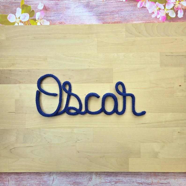 May include: A dark blue knitted cord shaped into the cursive name Oscar. The letters are formed from a single continuous piece of yarn with a textured, woven appearance, creating a clear and legible script style display of the name.