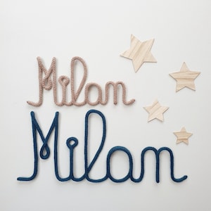 May include: Two names spelled Milan in cursive script, one crafted from light beige knitted yarn and one from dark blue knitted yarn, accompanied by four wooden star cutouts of varying sizes arranged to the right of the text.