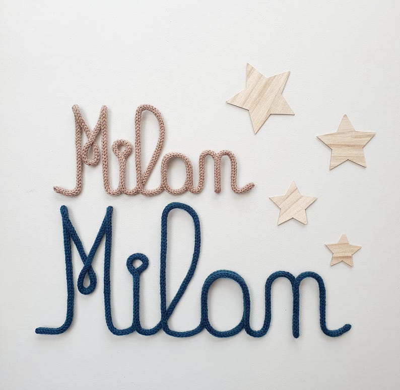 May include: Two names spelled Milan in cursive script, one crafted from light beige knitted yarn and one from dark blue knitted yarn, accompanied by four wooden star cutouts of varying sizes arranged to the right of the text.